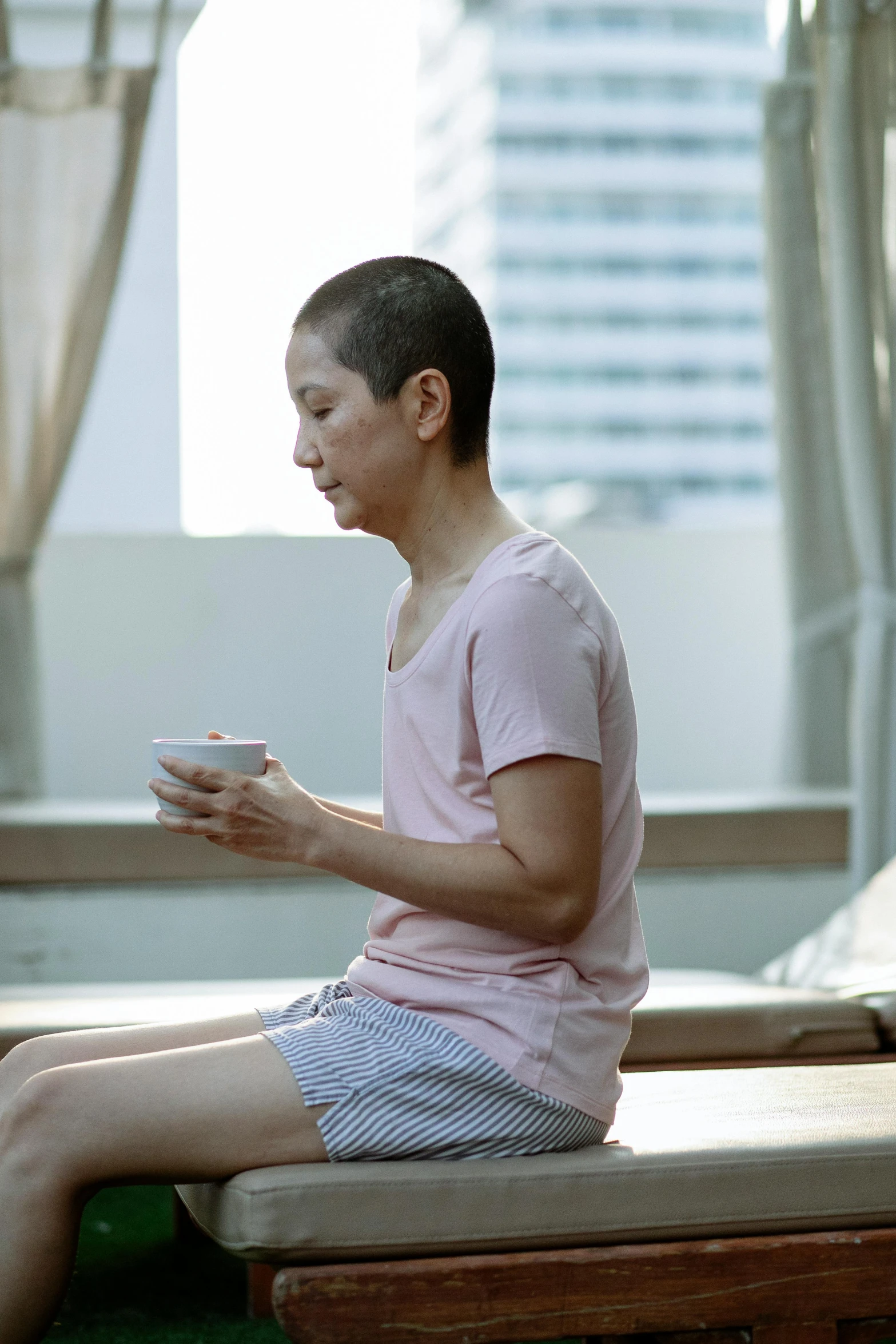 a woman sitting on a bed holding a cup, inspired by Fei Danxu, hyperrealism, partially bald, still from a live action movie, singapore, dance meditation