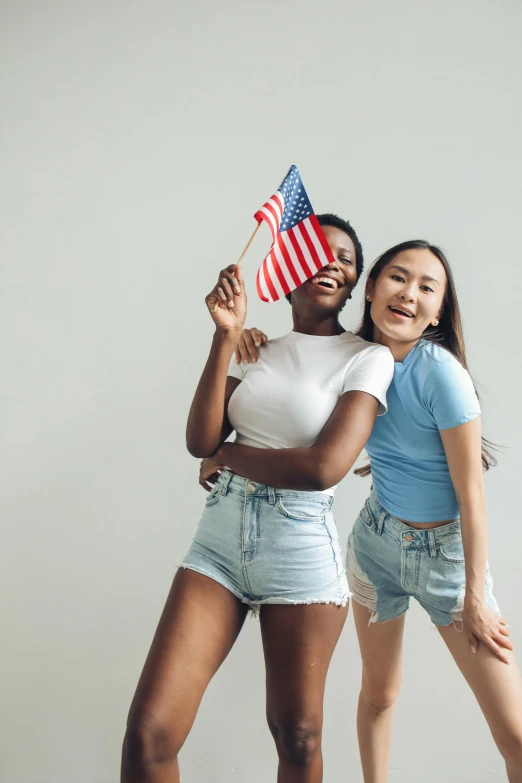 two women standing next to each other holding american flags, trending on unsplash, tan skin a tee shirt and shorts, on a gray background, tiktok, a young asian woman