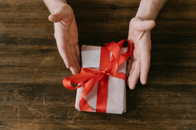 a person holding a gift wrapped in red ribbon, by Dan Content, pexels contest winner, arts and crafts movement, on a wooden desk, gif, manliness given form, packaging