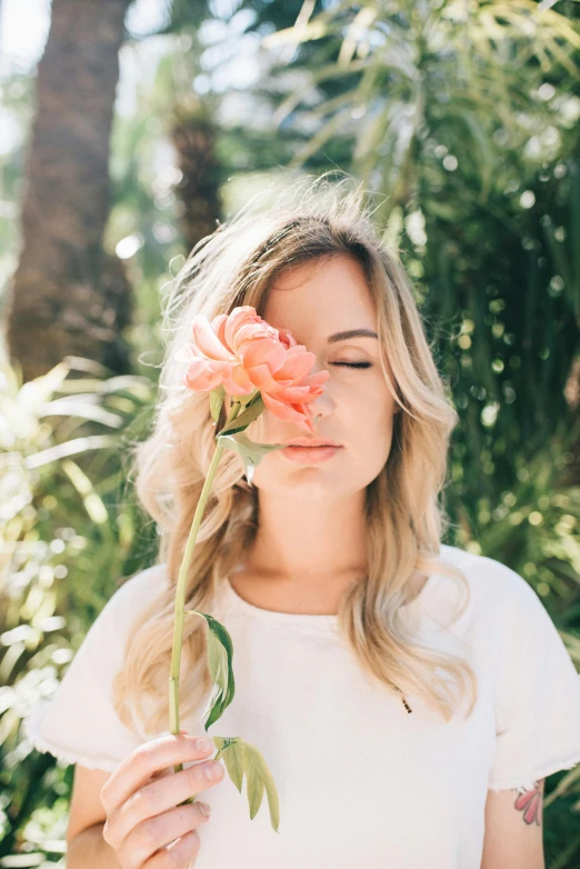 a woman holding a flower in front of her face, inspired by Károly Lotz, trending on unsplash, blonde flowing hair, lush surroundings, sydney sweeney, coral lipstick