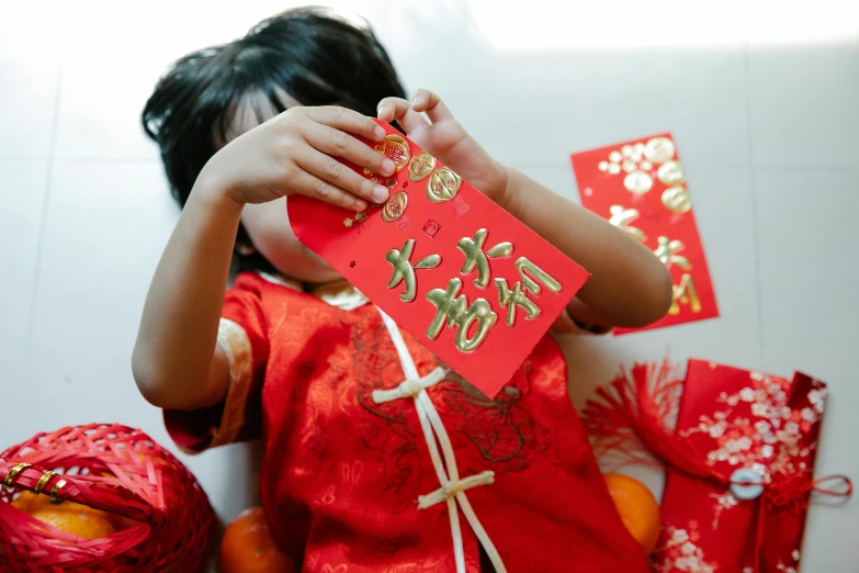 a little girl in a red dress holding a red envelope, by Julia Pishtar, pexels contest winner, golden chinese text, decorations, closeup of arms, celebration costume