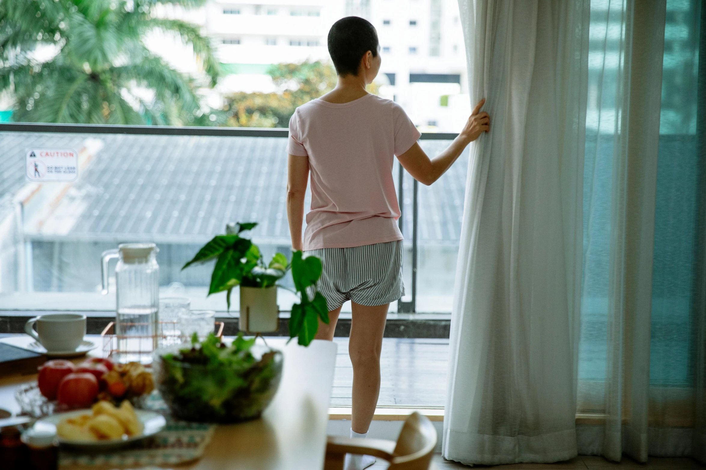 a woman standing in front of a window looking out, happening, wearing shorts and t shirt, waking up, gemma chen, curtains