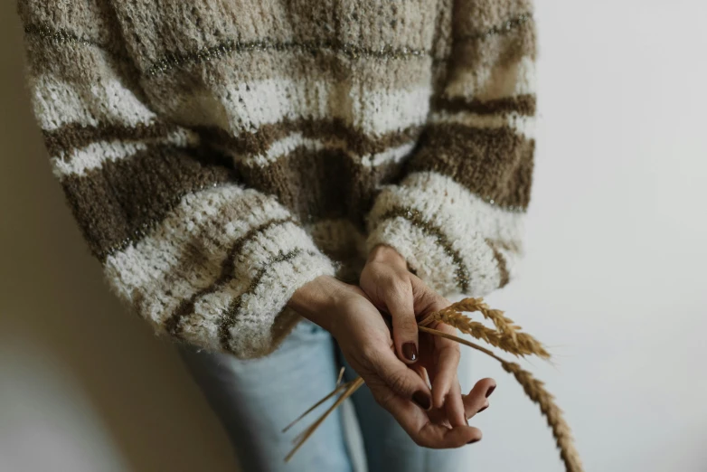 a woman holding a stalk of wheat in her hands, an album cover, by Emma Andijewska, trending on pexels, striped sweater, brown and white color scheme, texture, unedited