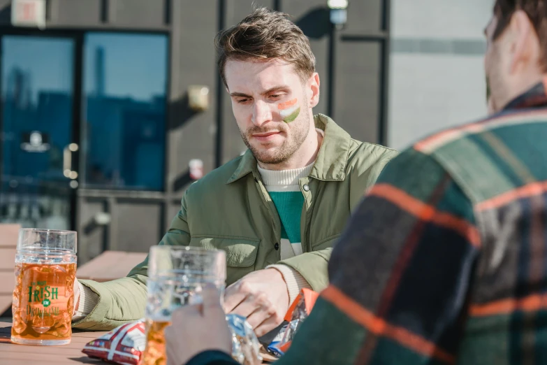 a man sitting at a table with a glass of beer, people outside eating meals, markings on his face, scobillyflup, rooftop party