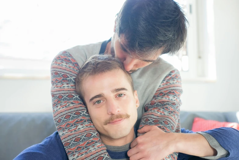 a man sitting on top of a couch next to a woman, a photo, two men hugging, light stubble, lachlan bailey, multicoloured