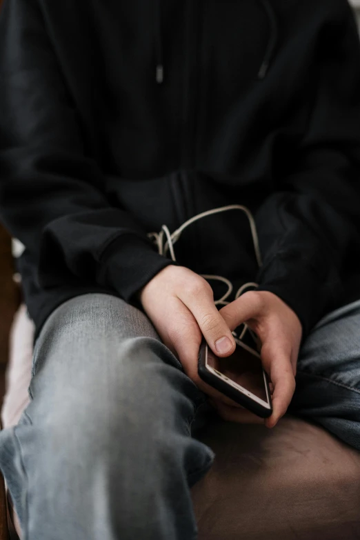 a close up of a person holding a cell phone, happening, wearing jeans and a black hoodie, cords and wires, sitting with wrists together, muted
