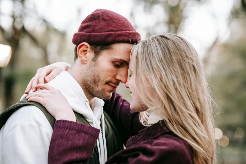 a man and a woman standing next to each other, trending on pexels, romanticism, maroon hat, cuddling, lachlan bailey, turning her head and smiling