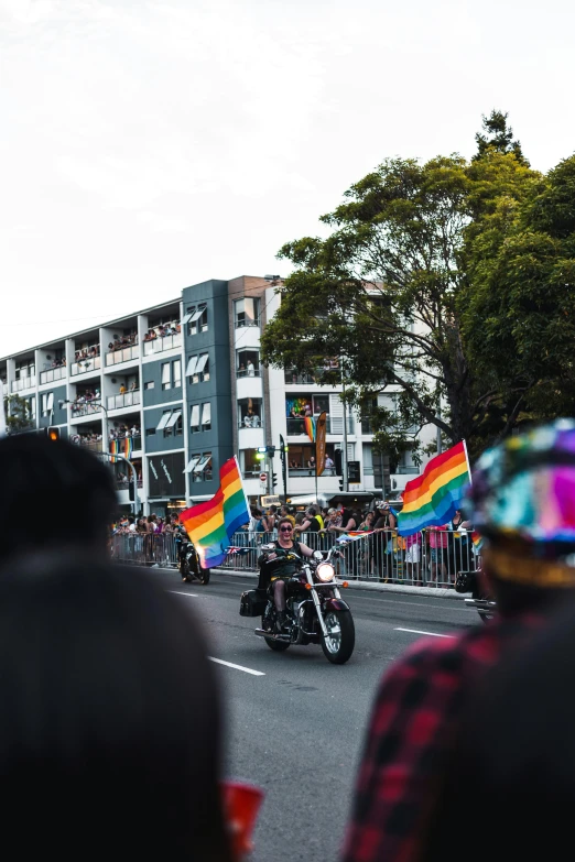 a group of people riding motorcycles down a street, pride flag in background, lachlan bailey, ultrawide lens”, fujifilm”