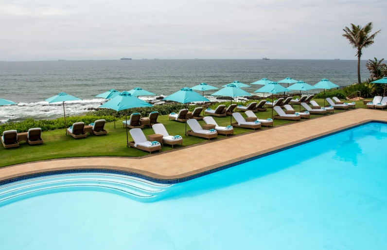 a pool with lounge chairs and umbrellas next to the ocean, zulu, sea foam, swimming pool