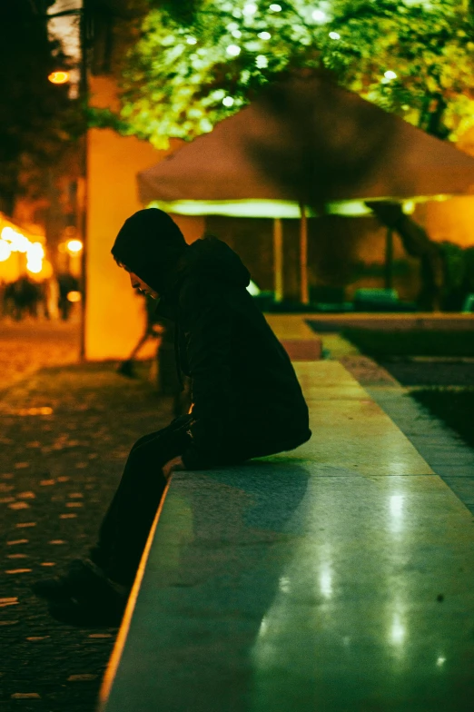 a person sitting on a bench at night, wearing jeans and a black hoodie, despondent, in a square, profile image