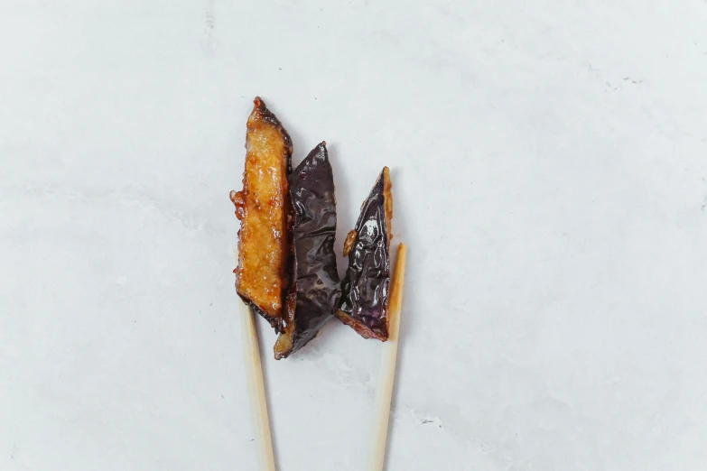 a couple of sticks that have some food on them, a picture, unsplash, mingei, eggplant, caramel, background image, on grey background
