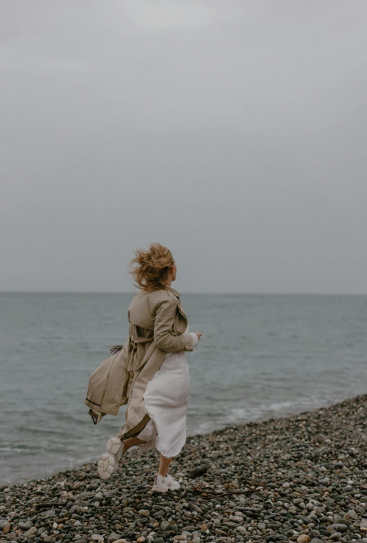 a woman walking on a rocky beach next to the ocean, pexels contest winner, renaissance, back of emma stone in beige coat, it's running between a storm, gif, dasha taran