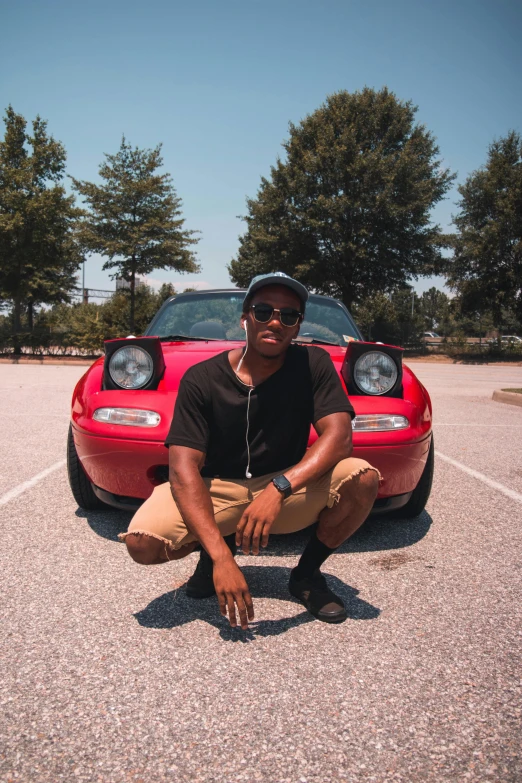 a man kneeling in front of a red sports car, an album cover, unsplash, photo in style of tyler mitchell, posing for a picture, vacation photo, low quality grainy