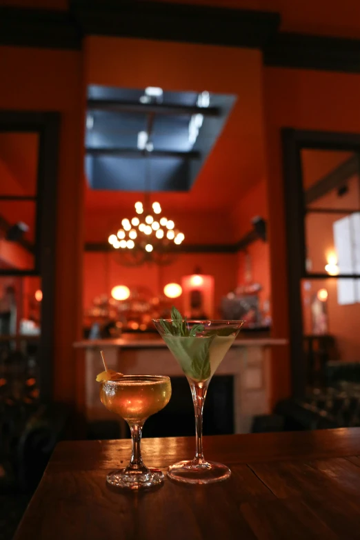 two cocktail glasses sitting on top of a wooden table, parlor, bustling, warm orange lighting, ivy's