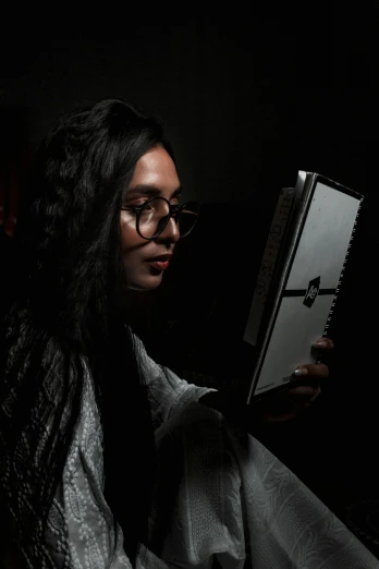 a woman reading a book in the dark, an album cover, by Sunil Das, pexels contest winner, bengal school of art, wavy long black hair and glasses, gamer, he is holding a large book, posed in profile