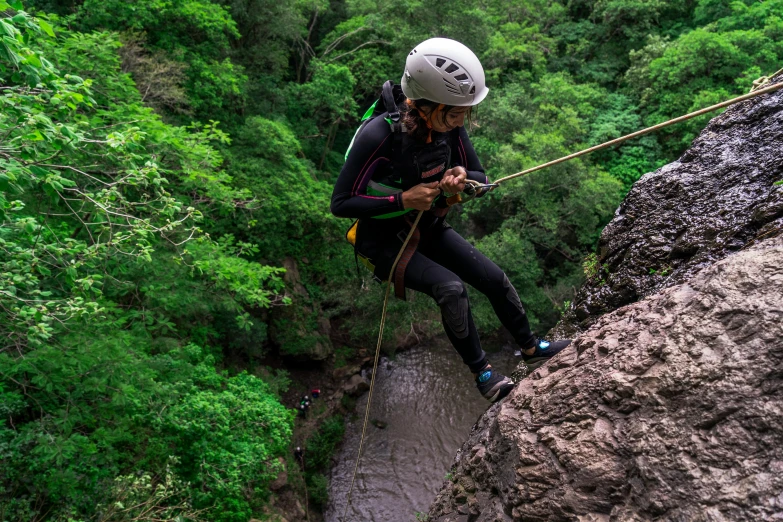 a man standing on top of a rock next to a river, hanging rope, helmet is off, lush green, avatar image