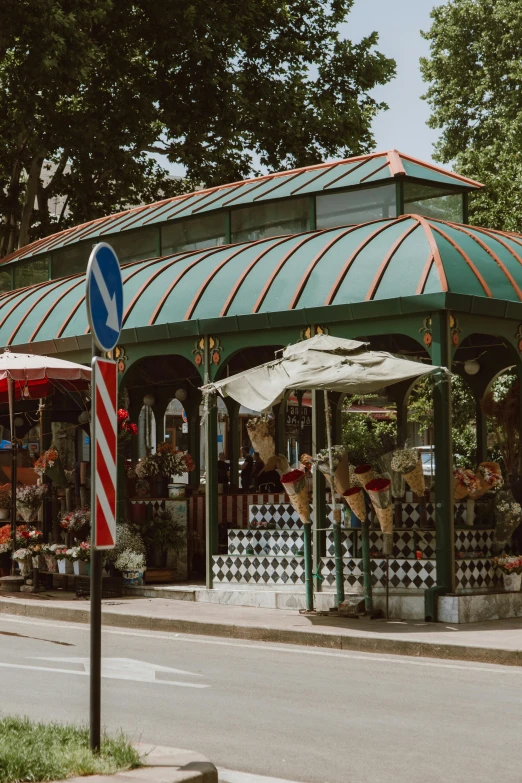 a couple of umbrellas sitting on the side of a road, with a french garden, colored market stand, lush trees and flowers, storefront