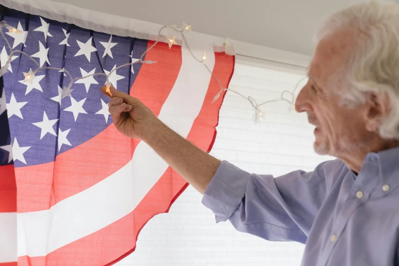 a man holding a string of lights in front of an american flag, an elderly, profile image, at home, with a white background