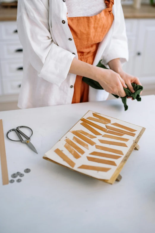 a woman in an apron preparing food on a table, inspired by Li Di, trending on pexels, arts and crafts movement, cut out of cardboard, eucalyptus, ribbon, set pieces