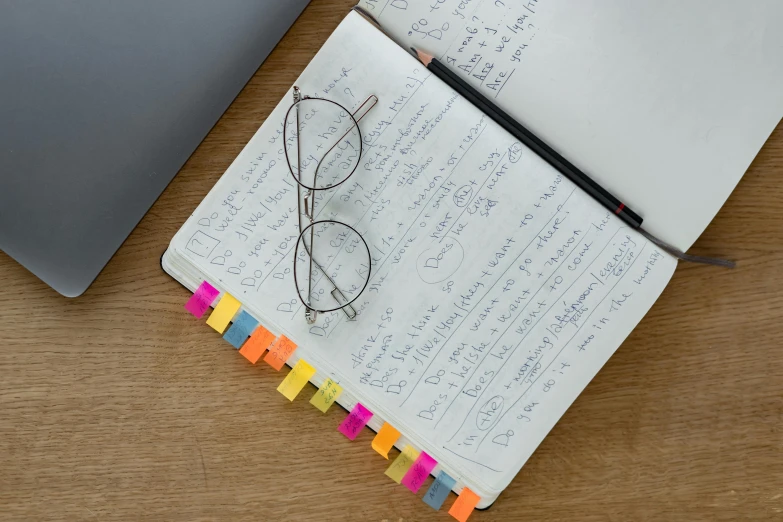 a notebook sitting on top of a wooden table next to a laptop, by Jessie Algie, unsplash, square rimmed glasses, rainbow neon strips, detailed plans and notes, detail shot