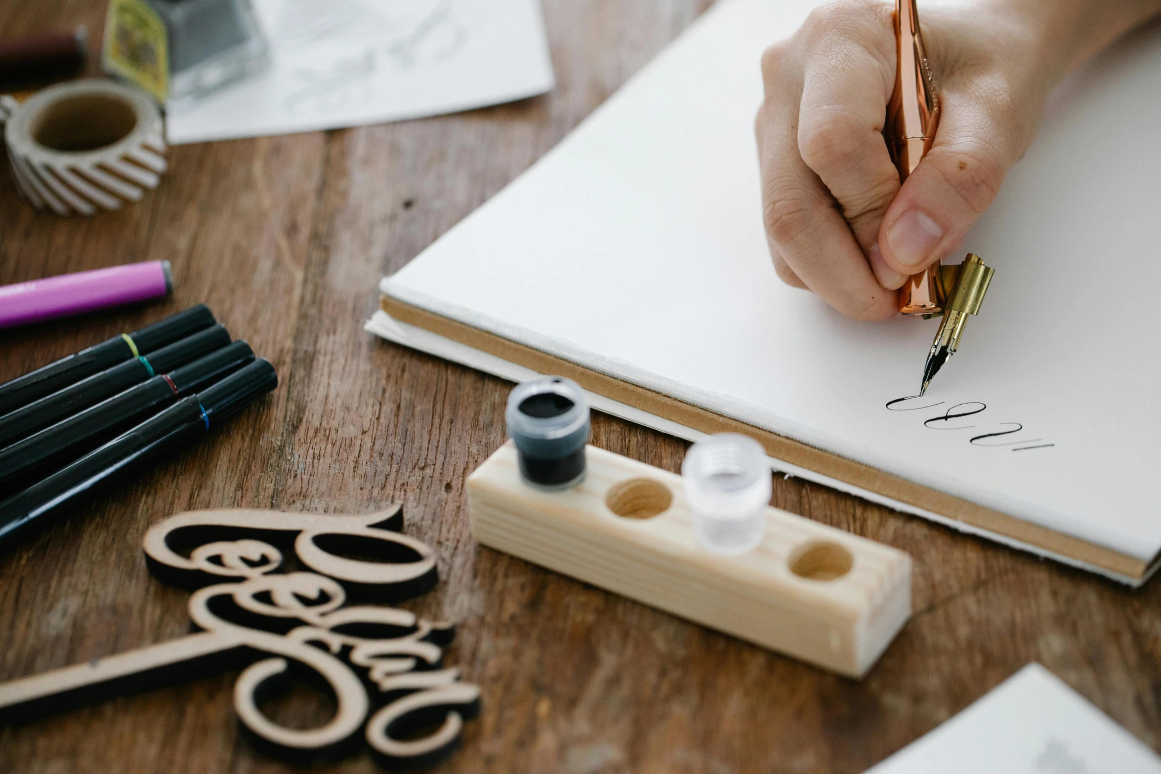 a person writing on a piece of paper with a pen, a drawing, trending on pexels, arbeitsrat für kunst, wooden art toys, “modern calligraphy art, 9 9 designs, painting on a badge