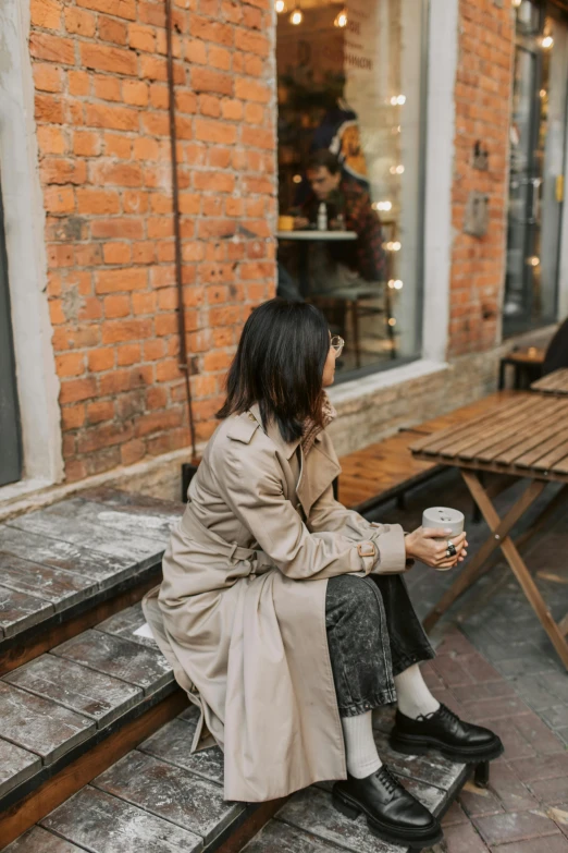 a woman sitting on a bench with a cup of coffee, trending on pexels, trench coat, louise zhang, cafe, muted browns