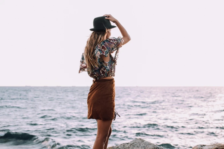 a woman standing on top of a rock next to the ocean, trending on pexels, renaissance, patterned clothing, wearing a fedora, wearing crop top and miniskirt, brown clothes