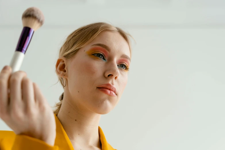 a woman holding a brush in front of her face, trending on pexels, hyperrealism, yellow makeup, looking across the shoulder, young blonde woman, half - length photo