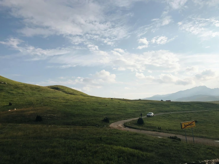 a car driving down a dirt road next to a lush green hillside, by Muggur, unsplash contest winner, les nabis, an expansive grassy plain, near lake baikal, neon signs in the distance, 2000s photo