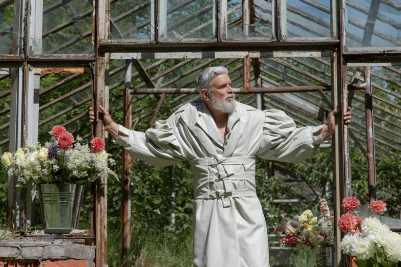 a man in a robe standing in front of a greenhouse, an album cover, inspired by Edwin Deakin, overalls and a white beard, ex machina, wearing silver silk robe, alchemical still made from clay