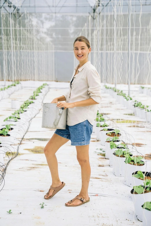 a woman standing in a greenhouse holding a watering can, a portrait, pexels contest winner, happening, wears shorts, hydroponic farms, wearing a linen shirt, in front of white back drop