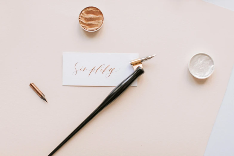 a pen sitting on top of a piece of paper next to a piece of paper with the word sunday written on it, an album cover, by Emma Andijewska, trending on pexels, letterism, rose gold, chinese ink brush, background image, on a pale background