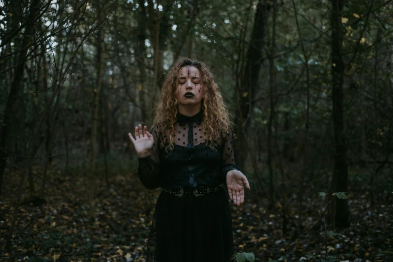 a woman in a black dress standing in a forest, an album cover, inspired by Elsa Bleda, pexels contest winner, gothic art, shrugging, portrait of a holy necromancer, home wicca scene, with haunted eyes and curly hair