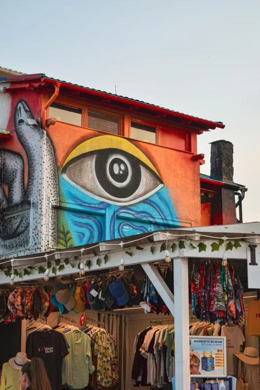 a store with a large eye painted on the side of it, trending on unsplash, street art, fishing village, psychedelic ski resort, next to the sea, indigenous art