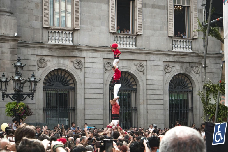 a group of people standing in front of a building, by Luis Molinari, pexels contest winner, figuration libre, acrobatic, cheering crowds, three towers, reds)