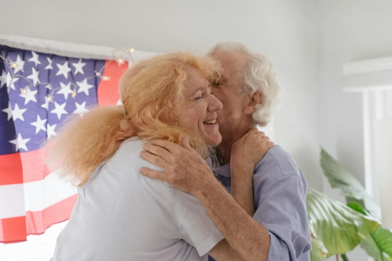 a man and woman hugging each other in front of an american flag, a colorized photo, by Harriet Zeitlin, trending on reddit, photorealism, an old man with a ginger beard, crazy white hair, nursing home, non-binary