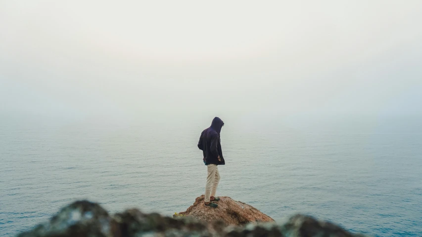 a person standing on top of a rock near the ocean, wearing a hoodie and sweatpants, overcast, profile image, distance fog