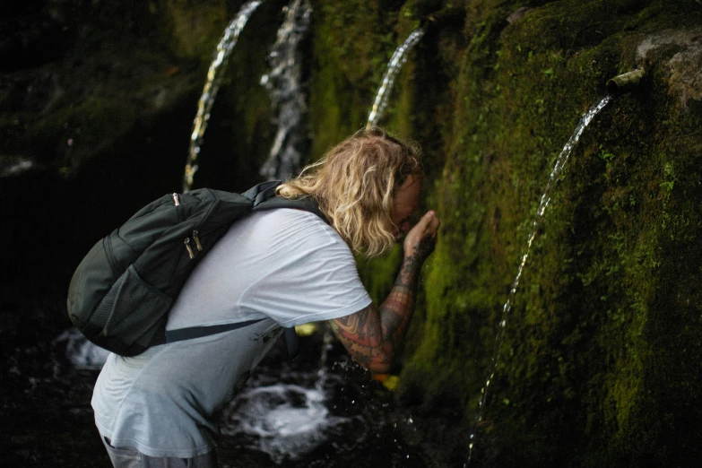 a man with a backpack standing in front of a waterfall, by Jesper Knudsen, graffiti, peacefully drinking river water, cottagecore hippie, profile image, covered in moss