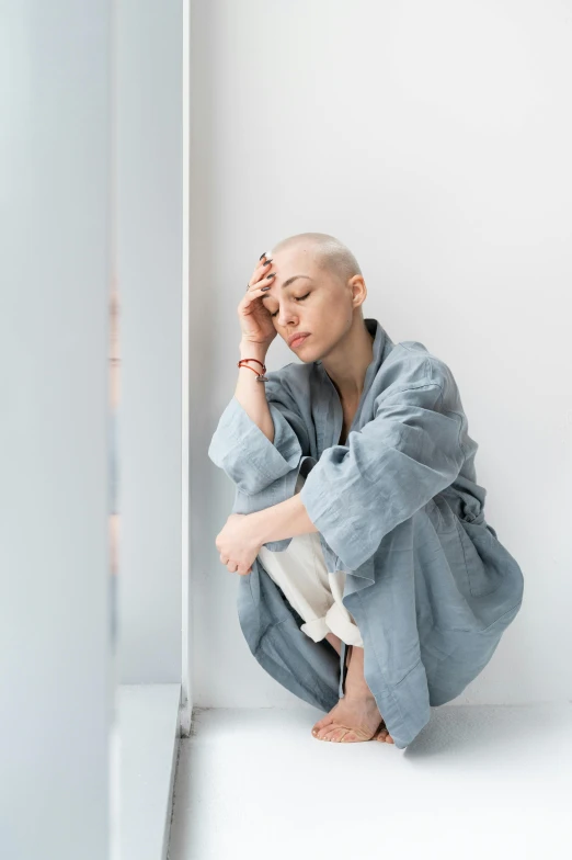 a woman sitting on the floor with her head in her hands, trending on unsplash, antipodeans, portrait of bald, bathrobe, blue gray, leaning on the wall