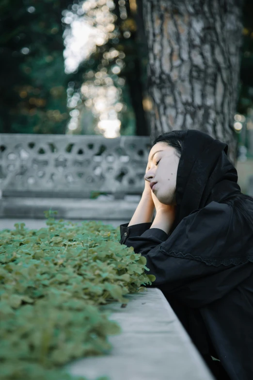 a woman sitting on a bench next to a tree, an album cover, unsplash, realism, wearing a dark hood, weeds and ivy on the graves, sleepy expression, asian woman