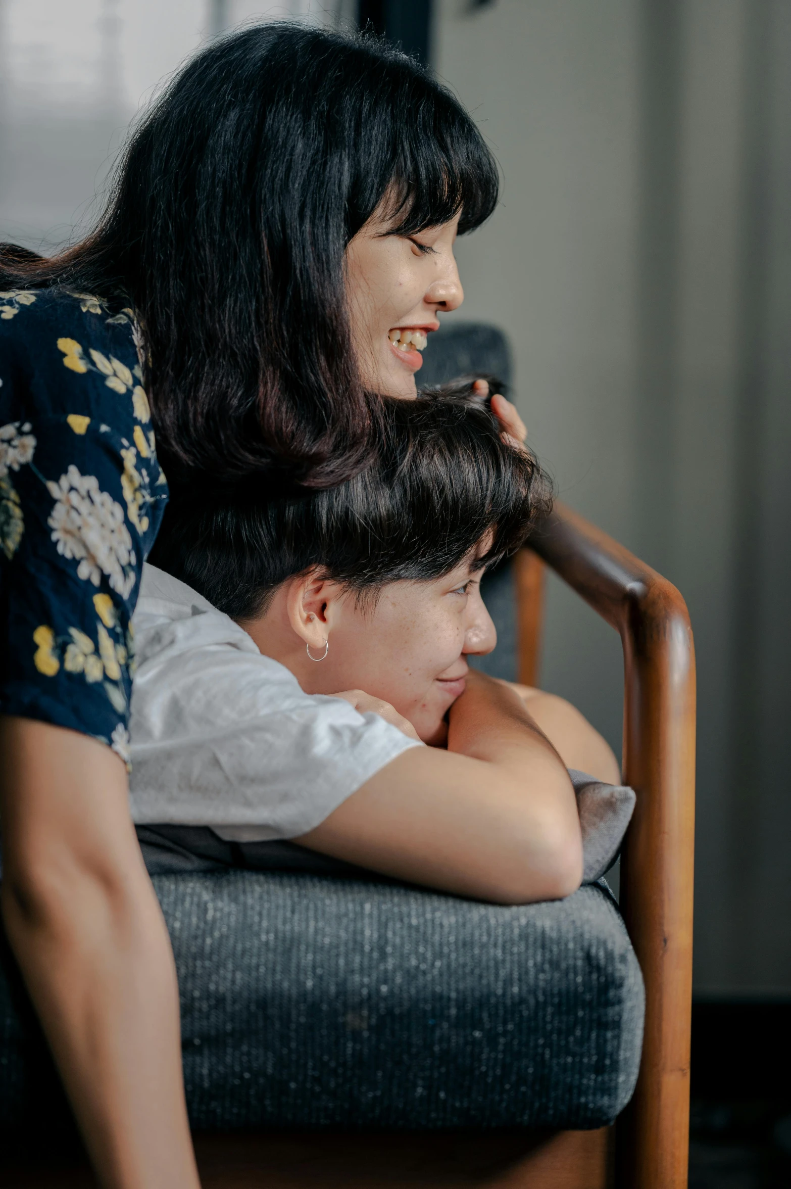 a woman sitting on top of a chair next to a boy, by Reuben Tam, pexels contest winner, incoherents, arm around her neck, profile image, relaxing on a couch, turning her head and smiling