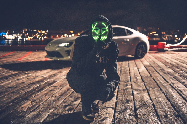 a person kneeling on a wooden floor with a car in the background, inspired by John Carpenter, pexels contest winner, elaborate lights. mask on face, green head, anonymous as a car, techwear