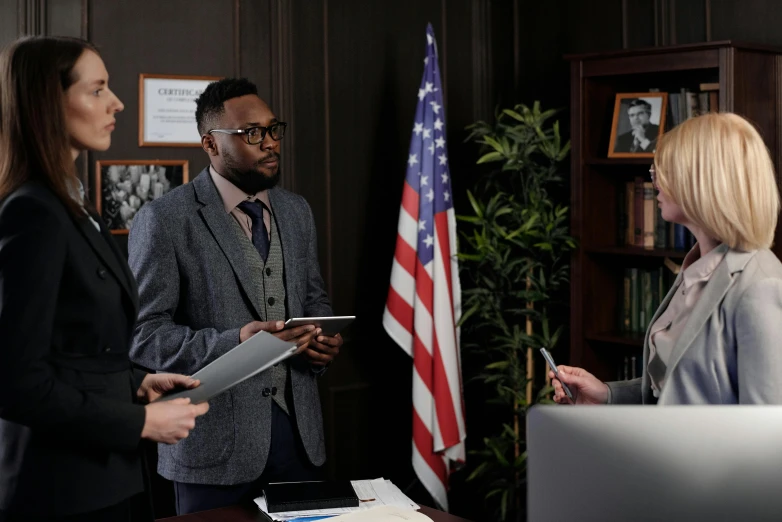 a group of people standing around a desk, his cape is the american flag, david uzochukwu, ( ( theatrical ) ), wearing a suit and glasses