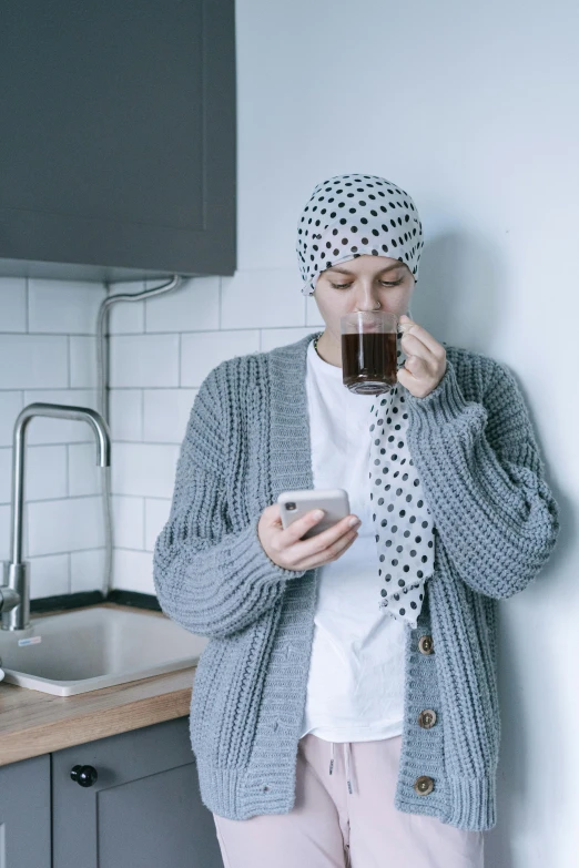a woman standing in a kitchen drinking a cup of coffee, inspired by Kun Can, hurufiyya, wearing beanie, genderless, sickness, checking her phone