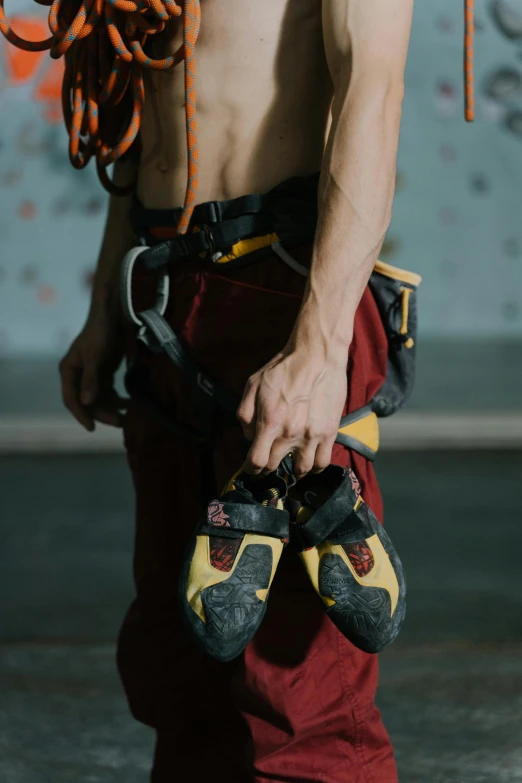 a shirtless man holding a pair of climbing shoes, pexels contest winner, renaissance, red and yellow, indoor shot, wearing adventure gear, cement