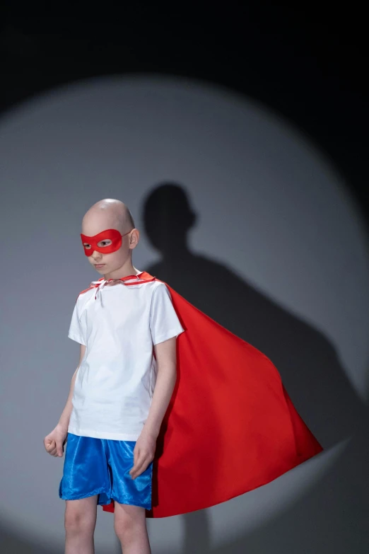 a young boy wearing a red cape and blue shorts, an album cover, shutterstock contest winner, antipodeans, bald head, super hero mask, fashion show photo, transgender