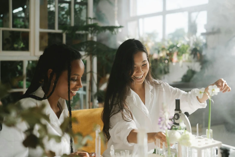 a couple of women sitting next to each other at a table, pexels contest winner, plants in beakers, wearing white silk robe, avatar image, bartending