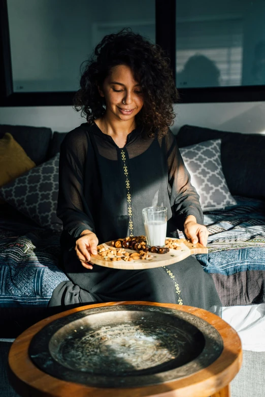 a woman sitting on a couch holding a plate of food, inspired by Riad Beyrouti, pexels, hurufiyya, black, grey, brown, morning glow