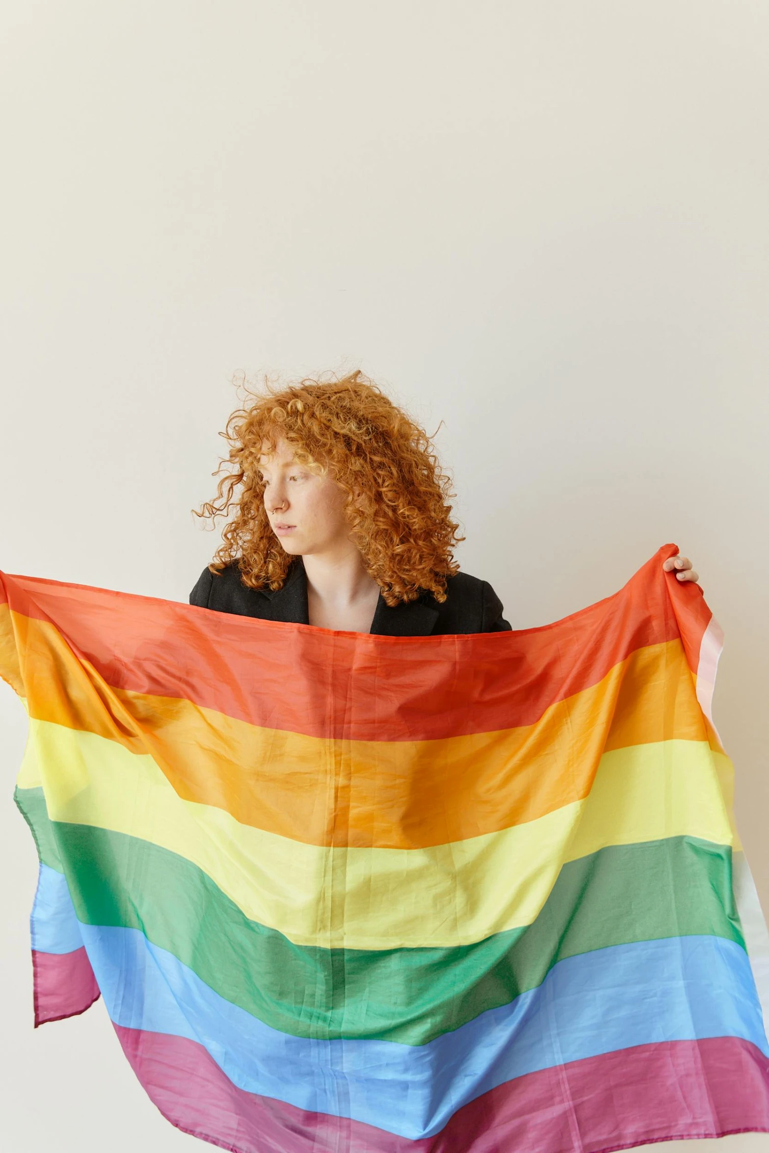 a woman holding up a rainbow colored blanket, by Emily Mason, with curly red hair, holding a white flag, on a pale background, ignant