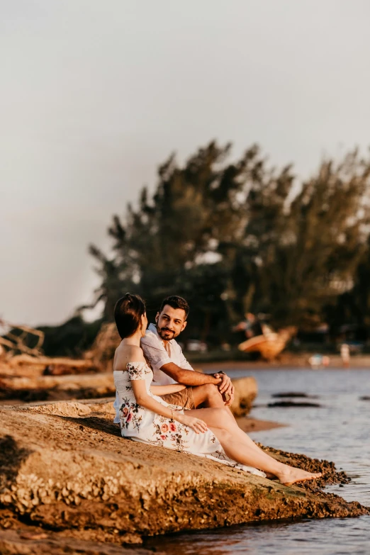 a man and woman sitting on a rock by the water, beach trees in the background, during golden hour, 2019 trending photo, puerto rico
